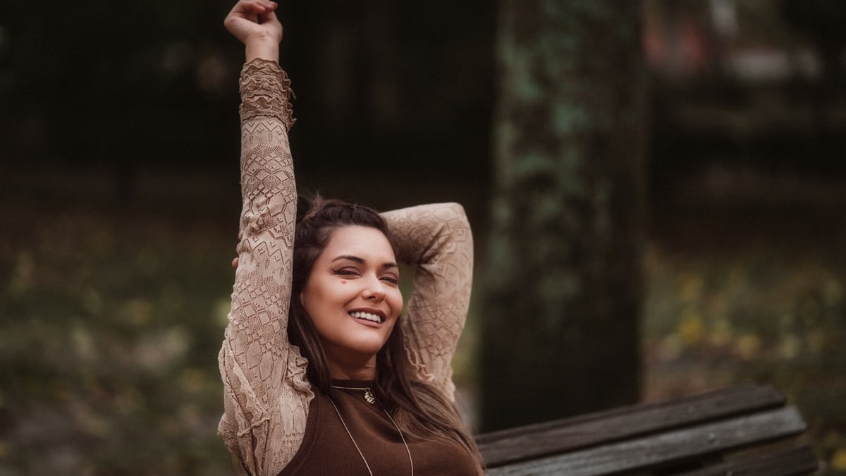 Smiling woman in brown outfit enjoying a peaceful autumn day in a Lisbon park, sitting on a wooden bench.